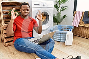 Young african man doing laundry and using computer relax and smiling with eyes closed doing meditation gesture with fingers