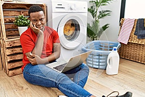 Young african man doing laundry and using computer thinking looking tired and bored with depression problems with crossed arms