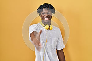 Young african man with dreadlocks standing over yellow background smiling friendly offering handshake as greeting and welcoming