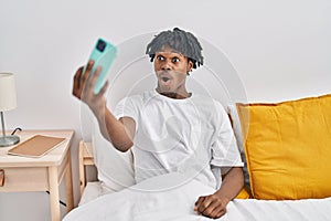 Young african man with dreadlocks taking a selfie on the bed scared and amazed with open mouth for surprise, disbelief face
