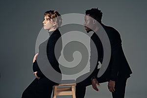 Young african man and his caucasian pregnant woman sit against grey background