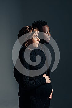 Young african man and his caucasian pregnant woman stand against grey background