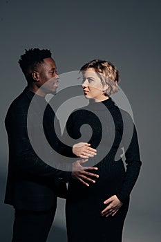 Young african man and his caucasian pregnant woman stand against grey background