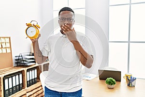 Young african man holding alarm clock at the office covering mouth with hand, shocked and afraid for mistake