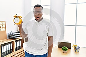 Young african man holding alarm clock at the office scared and amazed with open mouth for surprise, disbelief face
