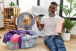 Young african man holding detergent bottle at laundry room smiling happy pointing with hand and finger to the side