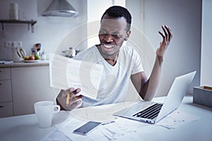 Young african man looking at bill, frustrated about high taxes, having problems with debt