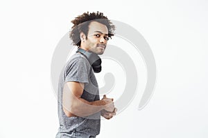 Young african man looking suspiciosly at camera standing in profile over white wall with his wireless headphones over