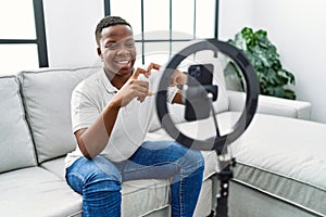 Young african man recording vlog tutorial with smartphone at home smiling in love showing heart symbol and shape with hands