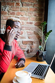 Young African man sitting at the coffee shop talk mobile phone. Student wearing trendy clothes, drinking coffee, using wireless In