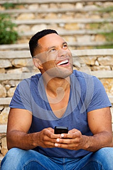 Young african man sitting outside using smart phone and looking up