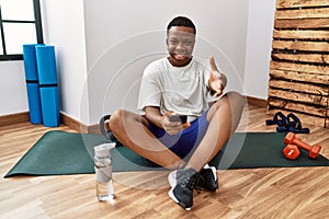 Young african man sitting on training mat at the gym using smartphone smiling friendly offering handshake as greeting and