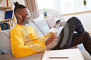 Young african man smiling, sitting on couch in earphones, looking in tablet, having online video call and drinking