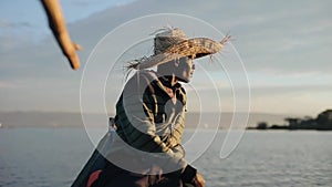 Young African man in straw hat sits in motor boat at dawn and look at sailing boats in sea.