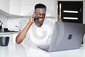 Young african man talk phone while use laptop in the kitchen