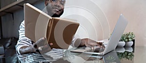 Young african man using laptop at home, black male looking at read book relaxing on leisure with work sit on glass table