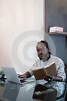 Young african man using laptop at home, black male looking at read book relaxing on leisure with work sit on glass table