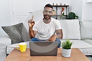 Young african man using laptop at home showing and pointing up with finger number one while smiling confident and happy