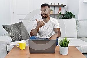 Young african man using laptop at home smiling with happy face looking and pointing to the side with thumb up
