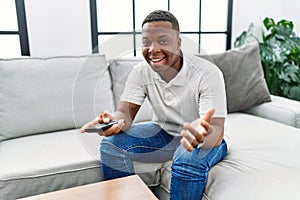 Young african man watching tv holding television remote control celebrating achievement with happy smile and winner expression