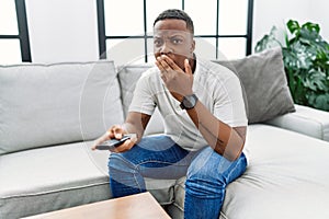 Young african man watching tv holding television remote control covering mouth with hand, shocked and afraid for mistake