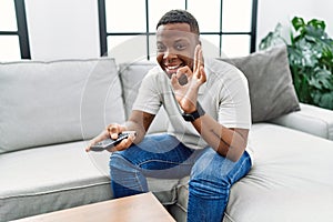 Young african man watching tv holding television remote control doing ok sign with fingers, smiling friendly gesturing excellent