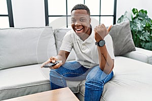 Young african man watching tv holding television remote control pointing thumb up to the side smiling happy with open mouth