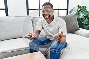 Young african man watching tv holding television remote control screaming proud, celebrating victory and success very excited with