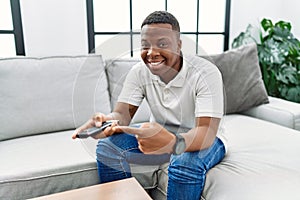 Young african man watching tv holding television remote control smiling happy pointing with hand and finger
