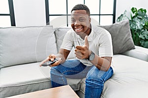 Young african man watching tv holding television remote control smiling happy and positive, thumb up doing excellent and approval