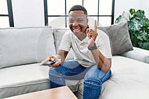 Young african man watching tv holding television remote control smiling with an idea or question pointing finger with happy face,