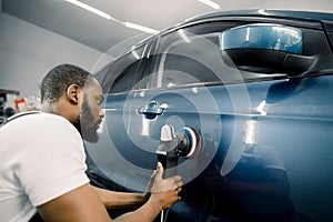 Young African man working at car detailing service, polishing blue car with polisher to eliminate contaminants from the