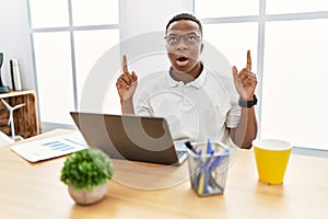 Young african man working at the office using computer laptop amazed and surprised looking up and pointing with fingers and raised