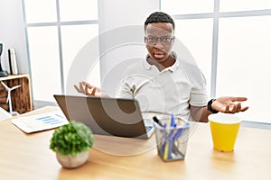 Young african man working at the office using computer laptop clueless and confused with open arms, no idea concept