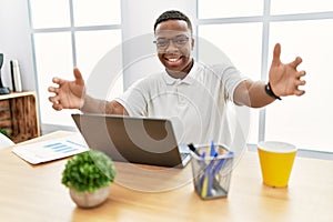 Young african man working at the office using computer laptop looking at the camera smiling with open arms for hug
