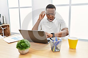 Young african man working at the office using computer laptop looking at the watch time worried, afraid of getting late