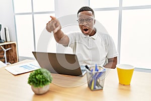 Young african man working at the office using computer laptop pointing with finger surprised ahead, open mouth amazed expression,