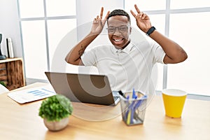 Young african man working at the office using computer laptop posing funny and crazy with fingers on head as bunny ears, smiling