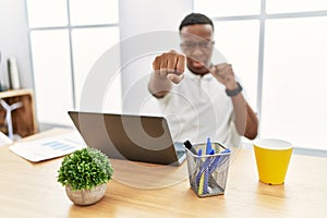 Young african man working at the office using computer laptop punching fist to fight, aggressive and angry attack, threat and