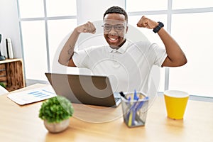 Young african man working at the office using computer laptop showing arms muscles smiling proud