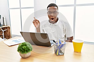 Young african man working at the office using computer laptop surprised pointing with finger to the side, open mouth amazed