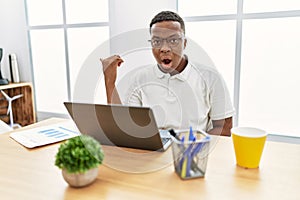 Young african man working at the office using computer laptop surprised pointing with hand finger to the side, open mouth amazed