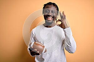 Young african player man playing rugby holding american football ball over yellow background doing ok sign with fingers, excellent