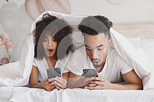 Young african shocked fun couple girl guy in t-shirts using mobile cell phone lying on bed with white sheet pillow