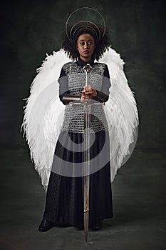 Young African woman with angel wings, halo above head and chainmail holding sword against vintage dark background.