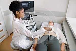 Young African woman doctor moving ultrasound transducer on woman`s belly in hospital. Female doctor giving patient ultra