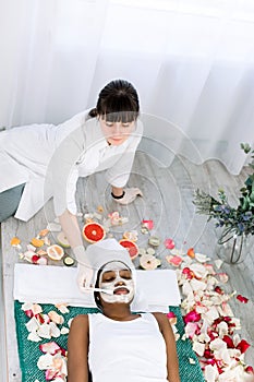 Young African Woman Getting Facial Mask At Spa, lying on the carpet on the floor with flowers. Natural cosmetics, skin