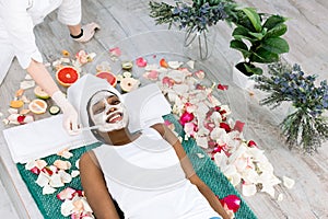 Young African Woman Getting Facial Mask At Spa, lying on the carpet on the floor with flowers. Natural cosmetics, skin