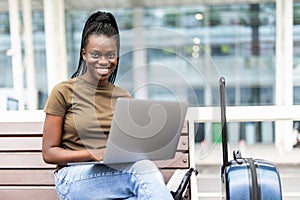 Young african woman with hand luggage in international airport terminal, working on her laptop while waiting for flight