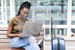 Young african woman with hand luggage in international airport terminal, working on her laptop while waiting for flight